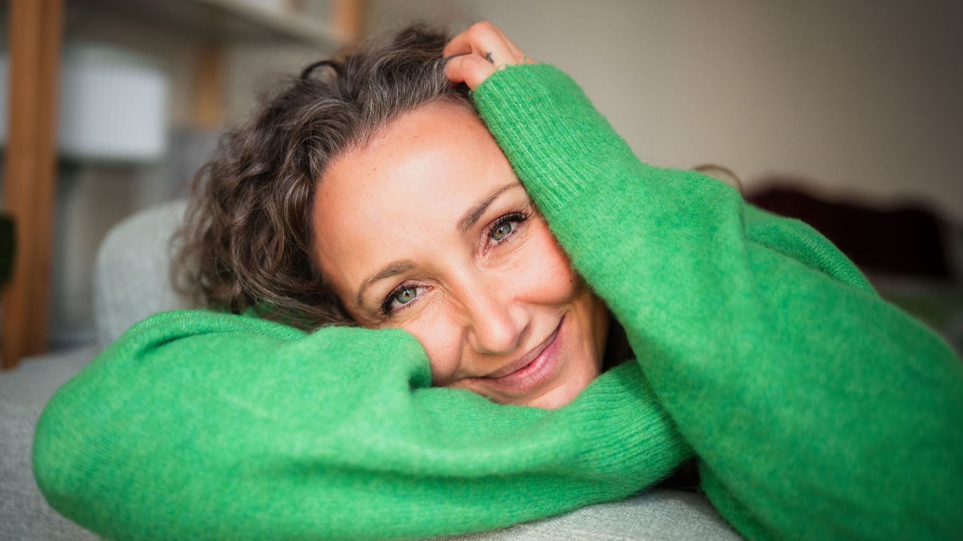 Mujer con suéter verde sonriendo, apoyando la cabeza en el brazo.